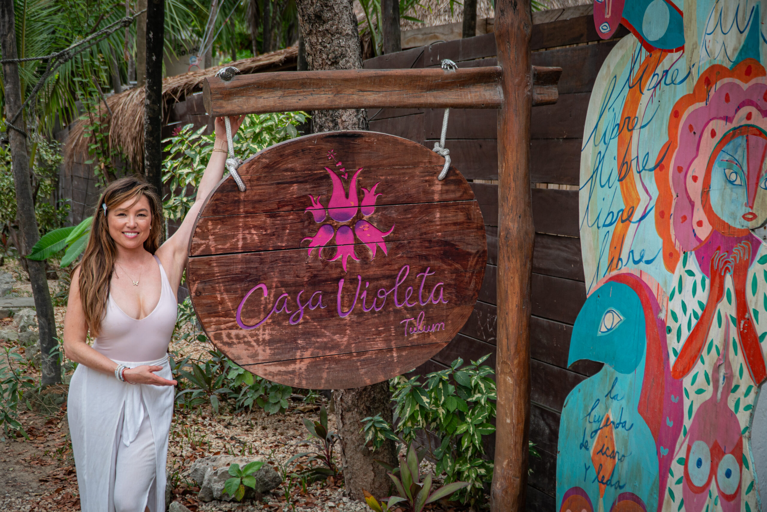 Woman standing beside a wooden sign that reads "Casa Violeta Tulum," a wellness retreat in Tulum, Mexico. She is smiling and wearing a white outfit, with tropical plants and a colorful mural in the background.