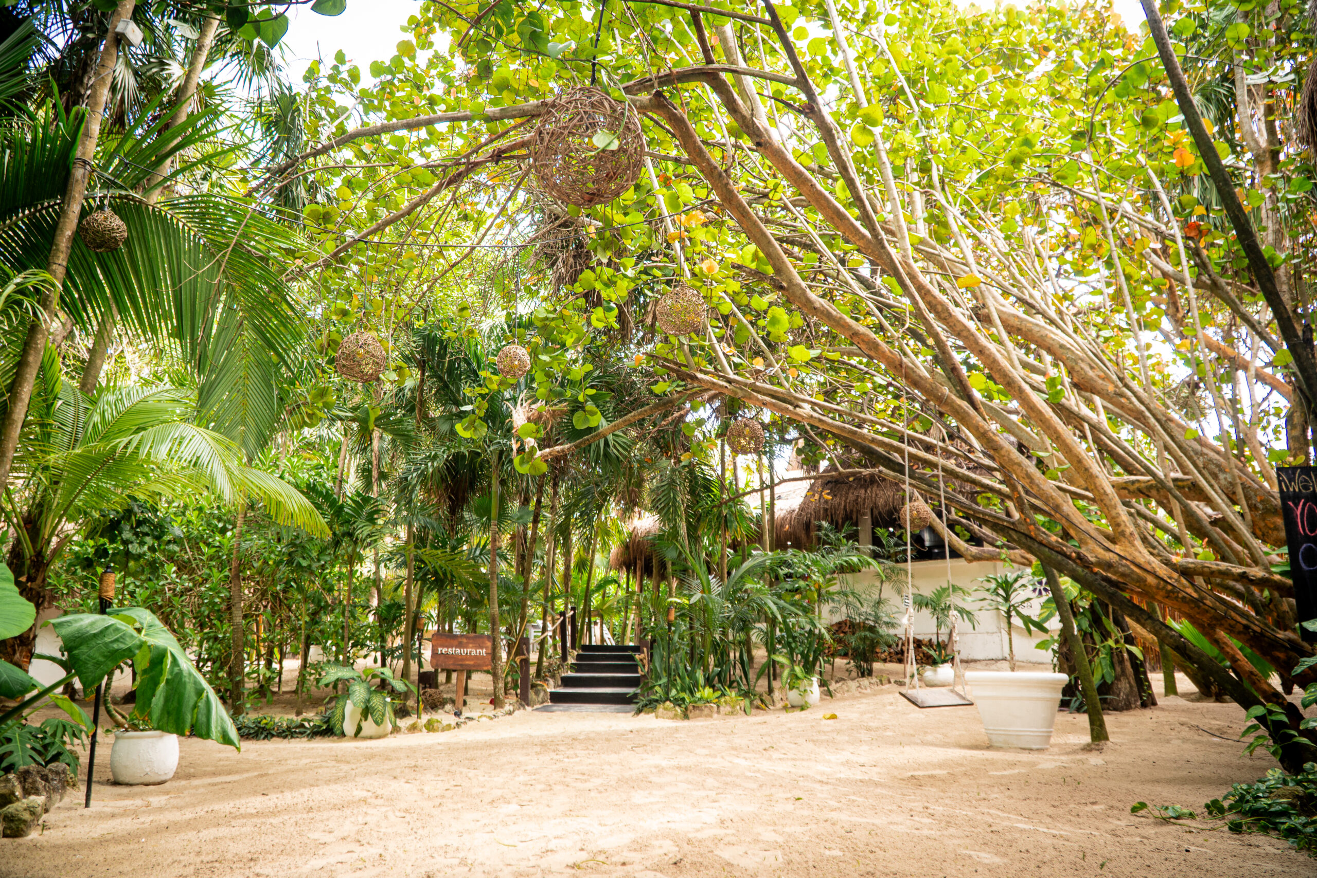 A serene tropical retreat with sandy paths, lush green foliage, hanging woven lanterns, and a wooden swing suspended from a tree. A small staircase leads to a thatched-roof structure, surrounded by vibrant plants.