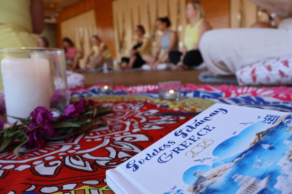 Close-up of a decorative altar with candle, flowers, and a Greece 2025 retreat journal at Cece Yoga’s Goddess Getaway, with participants seated in the background.