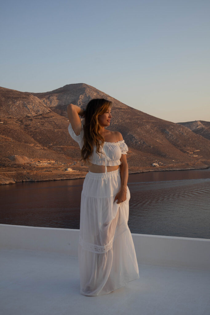 Woman in a white summer dress standing on a terrace with a view of mountains and sea at sunset in Greece.